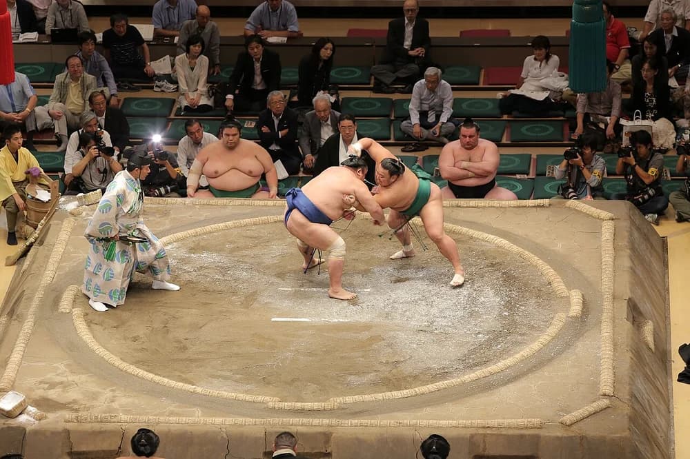 Two rikishi grappling in the middle of the dohyō, the gyōji to their side, mid-bout.