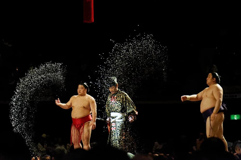 A rikishi tosses a great spray of salt across the dohyō during the shikiri ritual, the gyōji watching from centre.